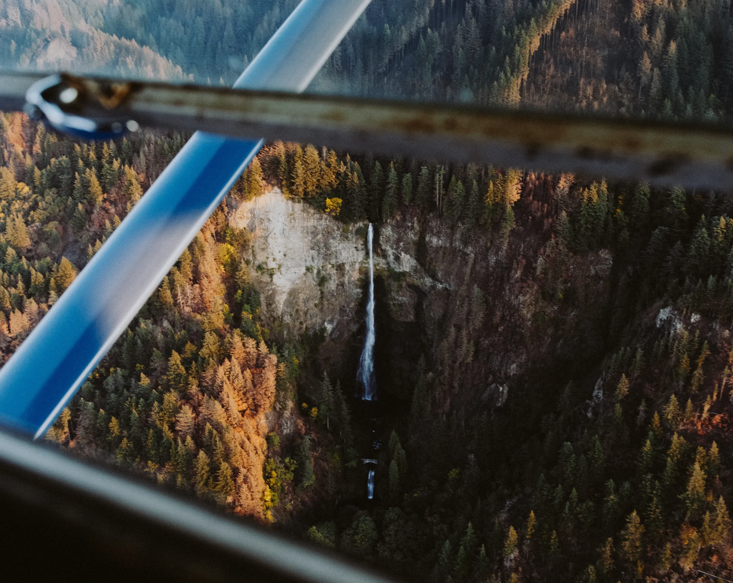 Aerial view of a waterfall cascading down a rocky cliff surrounded by dense, colorful forests in a scenic landscape.