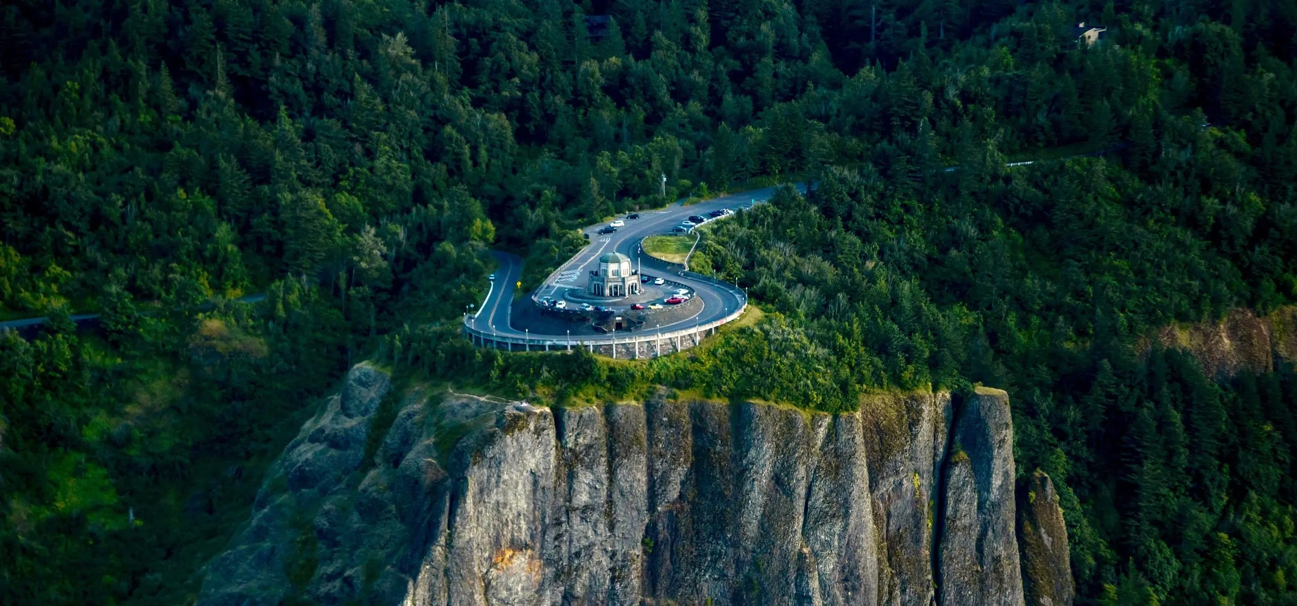 A scenic overlook surrounded by lush trees and a winding road, with a circular parking area atop a steep cliff.